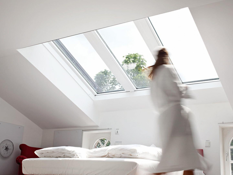 Unscharfe Frau im Bademantel läuft durch ein helles Schlafzimmer mit großem Panorama-Dachfenster